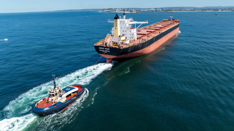 An electric tugboat and a large container ship on calm water on a sunny day
